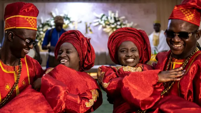Les frères jumeaux Kehinde et Taiwo Oguntoye portent Kehinde et Taiwo Adediran. Les deux couples portent des vêtements de cérémonie traditionnels rouges.